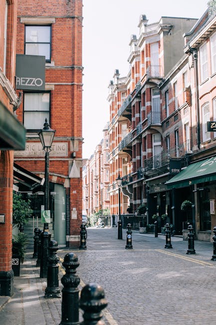 A quiet cobbled street in a residential and commercial area with Victorian-style brick buildings, including a prominent red-brick facade on the left with a sign for a business named 'HEZZO' visible on