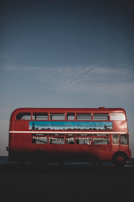 A red double-decker bus parked outdoors against a partly cloudy sky, featuring a large advertisement for the London Routemaster at Kensington Beach on the side. The bus is positioned on a paved surfac